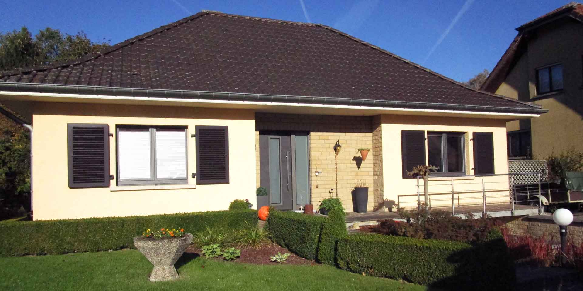 Gelbliches Einfamilienhaus mit einer modernen grauen Coplaning Keramik Haustür und braunen Coplaning Holz Jalousienfenster.