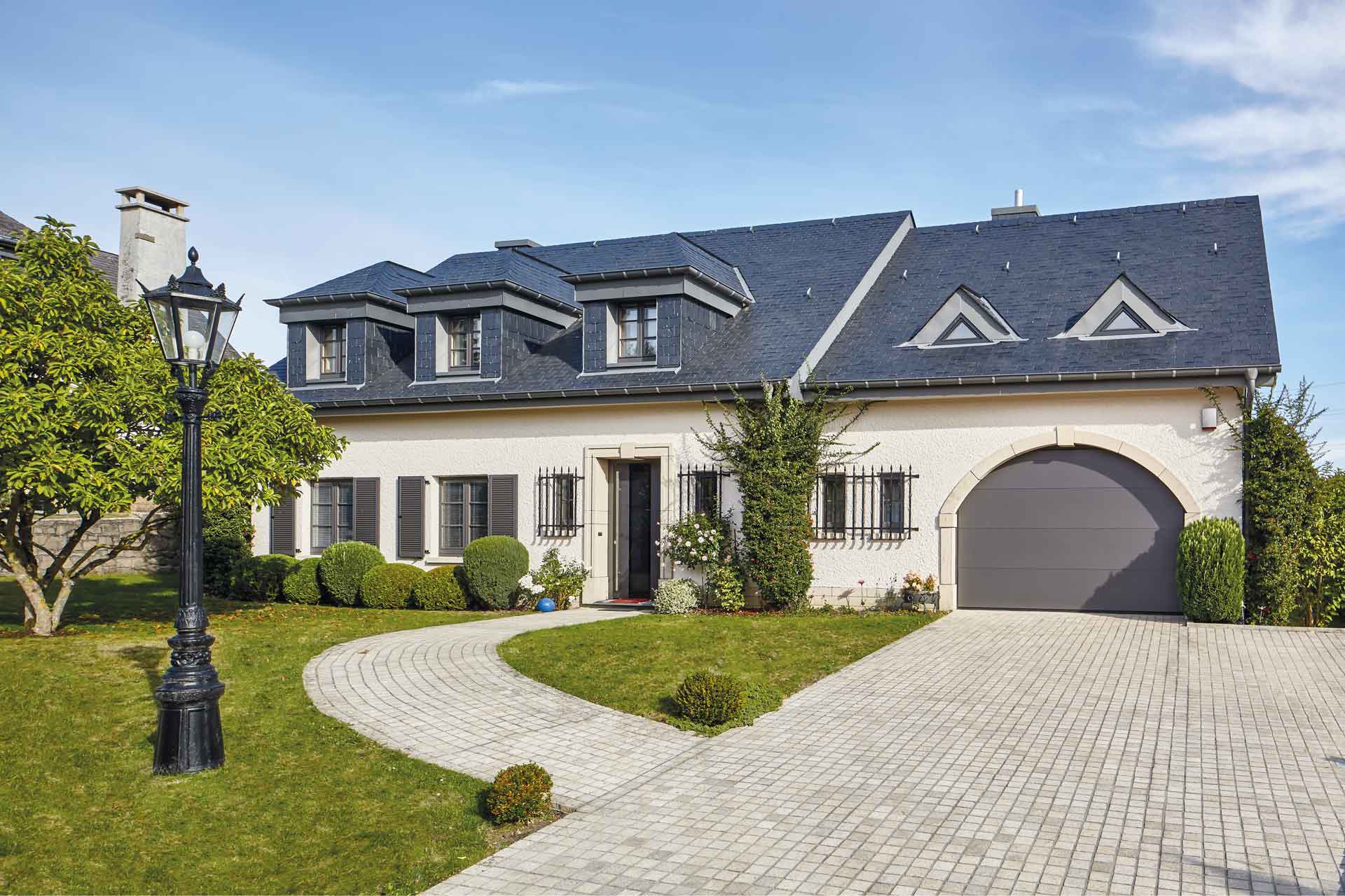 Weißes Einfamilienhaus mit grauer Coplaning Keramik Haustür, braunen Coplaning Holz- Alu Fenster und einem grauen Deckensektionalen Garagentor.