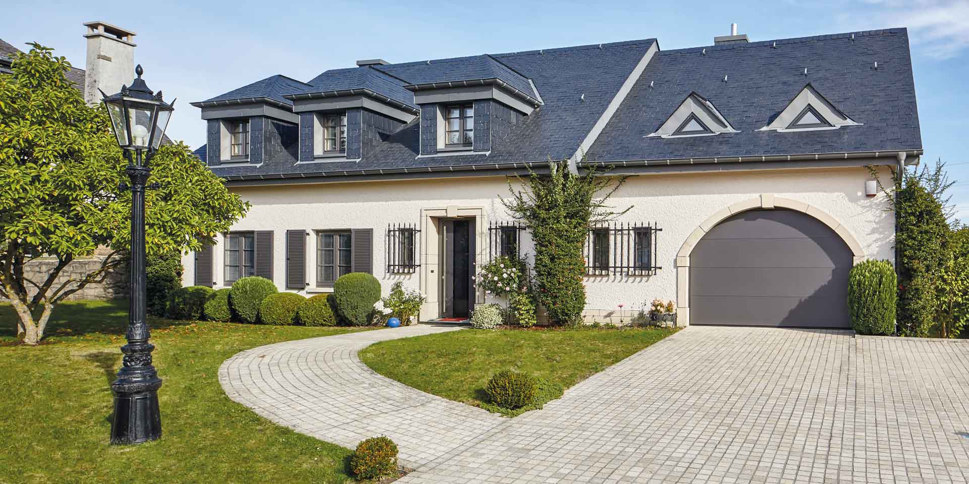 Weißes Einfamilienhaus mit grauer Coplaning Keramik Haustür, braunen Coplaning Holz- Alu Fenster und einem grauen Deckensektionalen Garagentor.