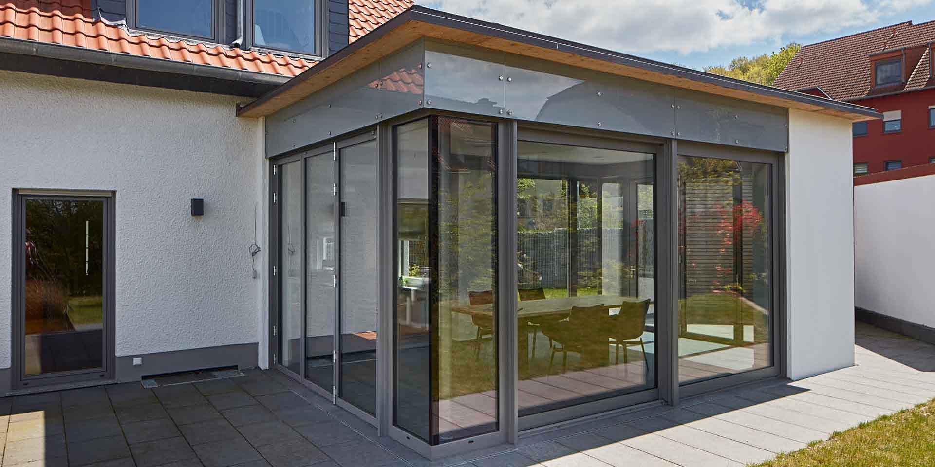 Weißes Einfamilienhaus mit einem grauen Aluminium Coplaning Veranda Anbau mit Faltanlage, Glasfaltwand und Ganzglasecken.