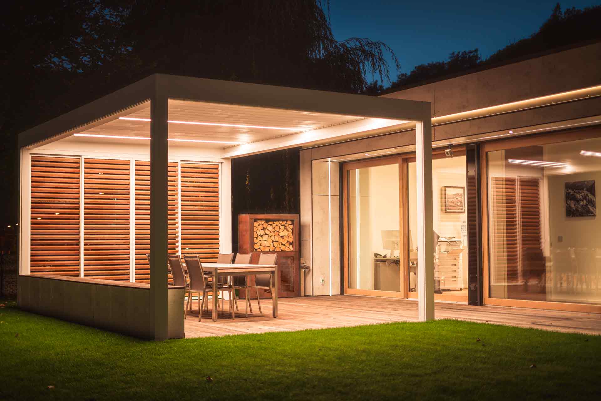 Modernes Einfamilienhaus mit einem hochwertigen Coplaning Pergola in der Nacht mit moderner Terassenüberdachung eind einem Quadrato in weiß mit Lamellendach.