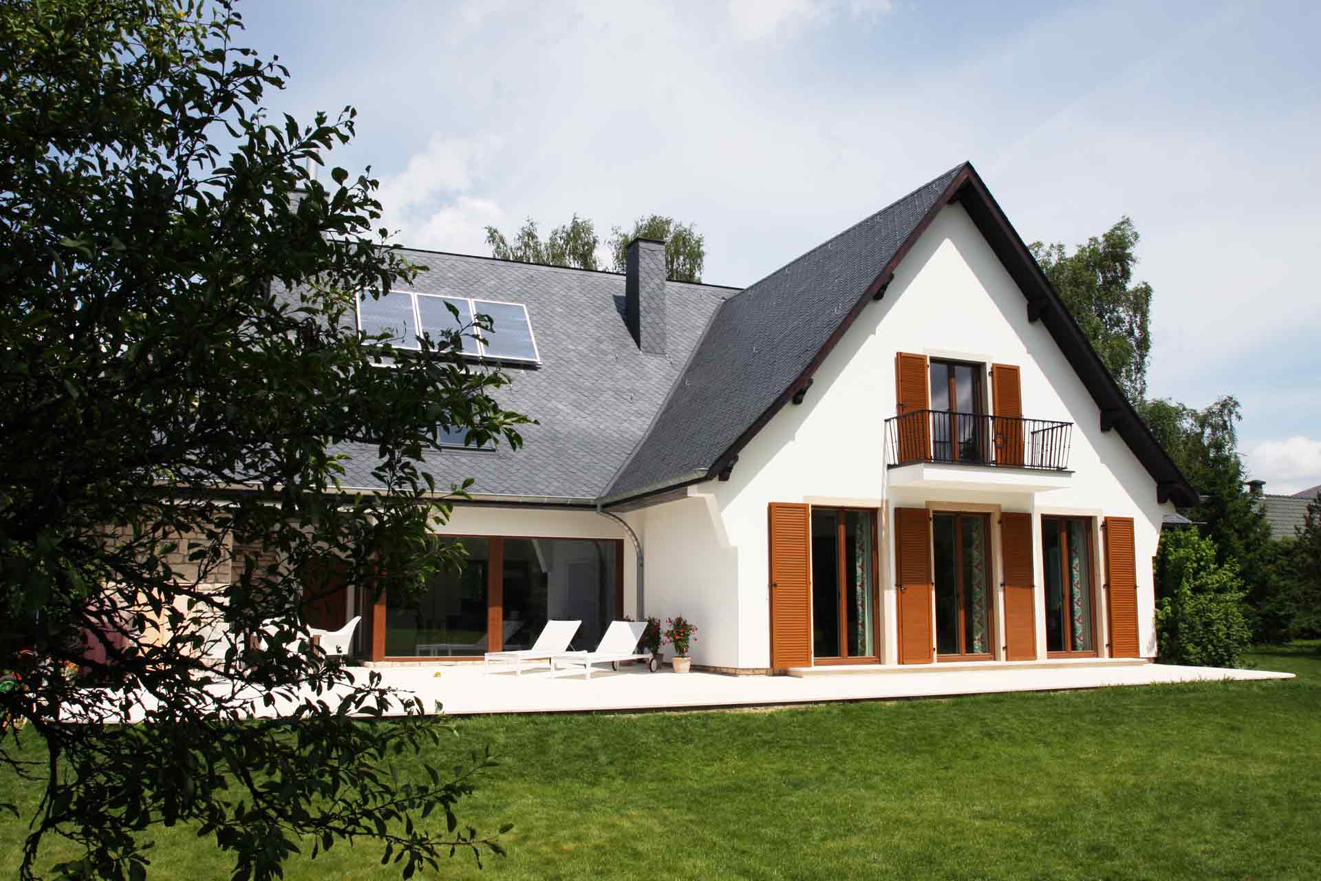 Rückseite von links gesehen man sieht ein großes weißes Haus mit Coplaning Holzfenster und Fenstertüren mit Klappläden aus Holz mit integrierten Jalousien.