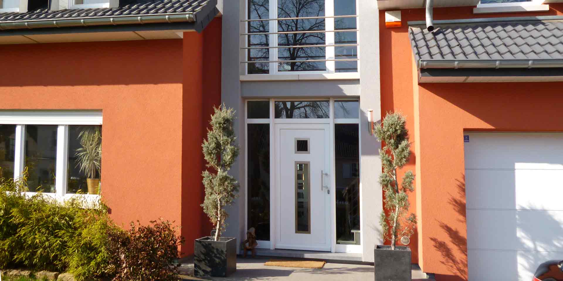 Nahaufnahme des Eingangsbereichs eines roten Einfamilienhauses mit Blick auf eine weiße moderne Coplaning Haustür mit Glas und darüber ein großes weißes Coplaning Gauben- Kunststoff Fenster.