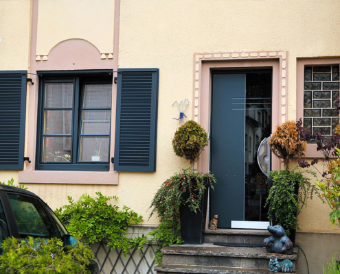 Gelbes Einfamilienhaus mit Nahaufnahme des Eingangsbereichs mit Blick auf eine graue Coplaning Aluminium Haustür und grauen Coplaning Kunststoff Fenster mit grauen Aluminium Klappläden.