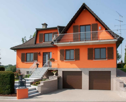 Rotes Einfamilienhaus mit grau- brauner Coplaning Haustür und braunen Coplaning Fenster mit Klappläden und zwei braunen Garagentor.