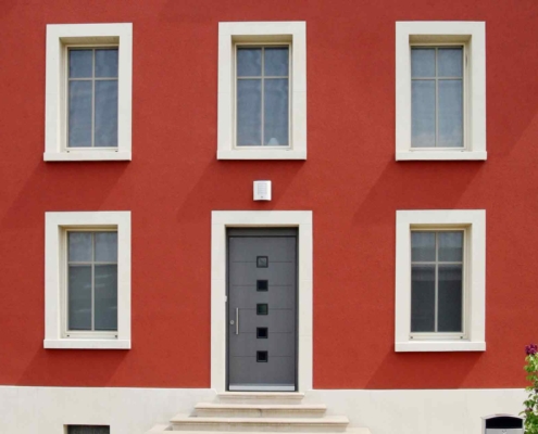 Nahaufnahme der Vorderseite des roten Einfamilienhauses mit grauer Coplaning Haustür mit Glasauschnitten und mit weißen Coplaning Fenster.