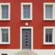 Nahaufnahme der Vorderseite des roten Einfamilienhauses mit grauer Coplaning Haustür mit Glasauschnitten und mit weißen Coplaning Fenster.