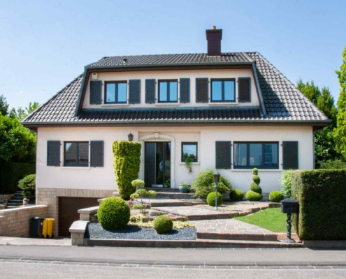 Leicht pinkes Einfamilienhaus mit moderner grau- schwarzen Coplaning Haustür,grauen Coplaning Fenster mit grauen Klappläden und mit einem Garagentor.