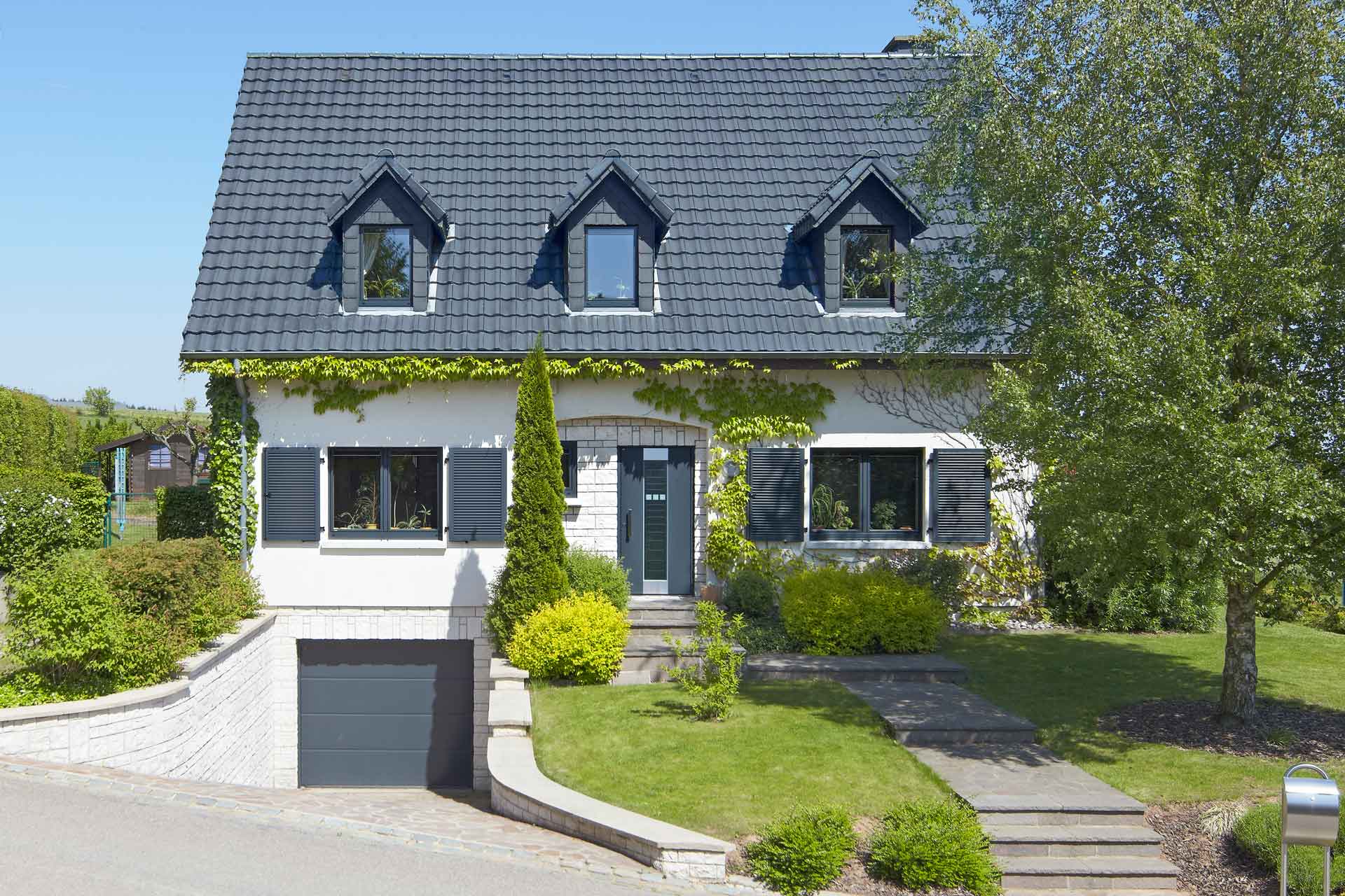 Weißes Einfamilienhaus mit neuen modernen Haustür und grauen einbruchsicheren Coplaning Fenster und einem grauen Garagentor.