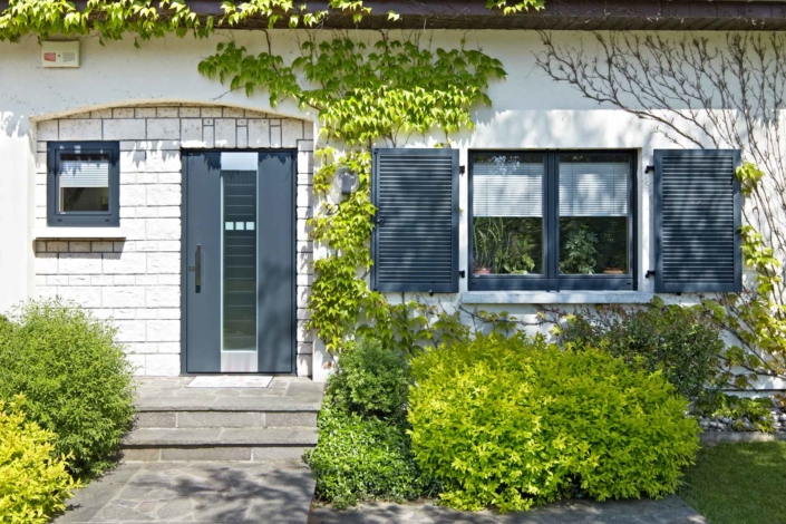 Nahaufnahme des Eingangsbereichs eines weißen Einfamilienhauses mit Blick auf die neue moderne graue Coplaning Haustür und jeweils links und rechts davon ein graues einbruchsicheres Coplaning Fenster.