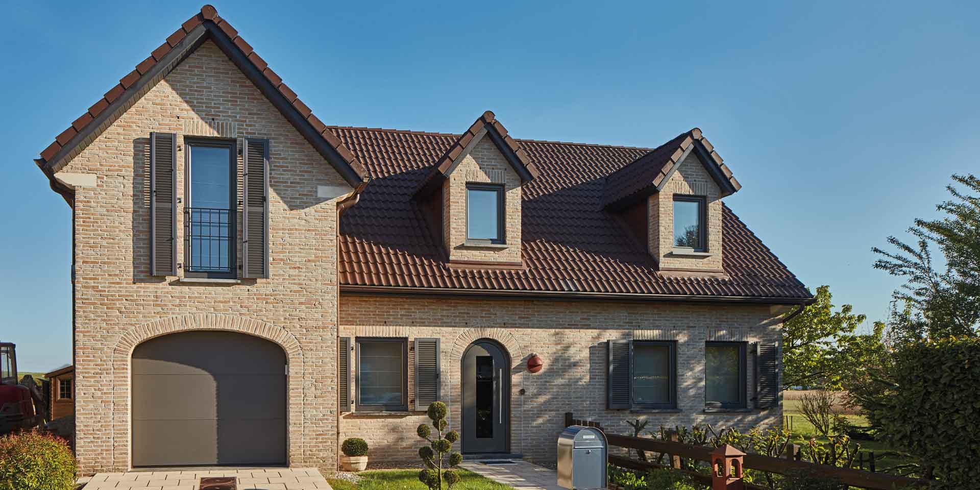 Einfamilienhaus mit Steinfassade mit neuer grauen runden Coplaning Alu Haustür mit grauen Coplaning Holz- Alu Fenster und einem grauen Garagentor.