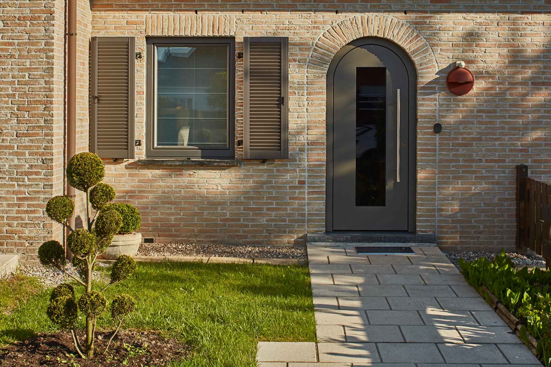 Nahaufnahme des Eingangsbereich eines Einfamilienhauses mit Steinfassade mit einer neuen runden Coplaning Haustür und links davon ein graues Coplaning Alu Fenster.