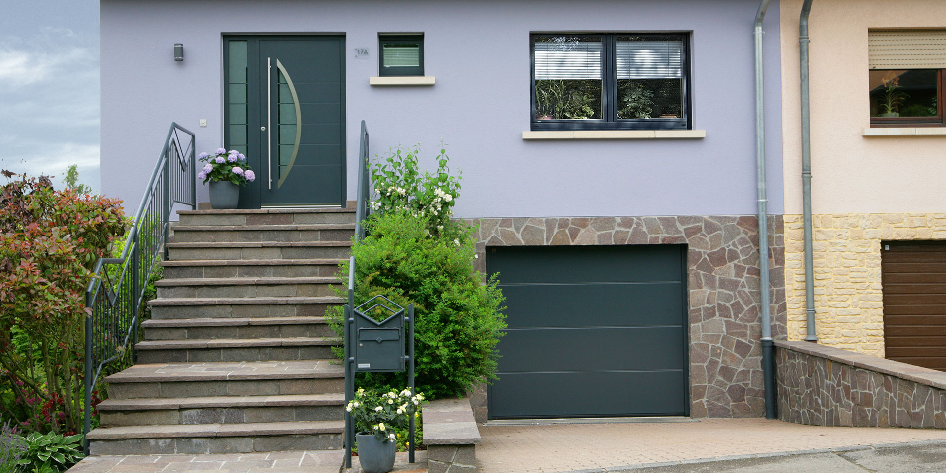 Lila Einfamilienhaus mit einer grau lackierten Coplaning Haustür, grauen Coplaning Fenster und einem grauen Coplaning Garagentor.