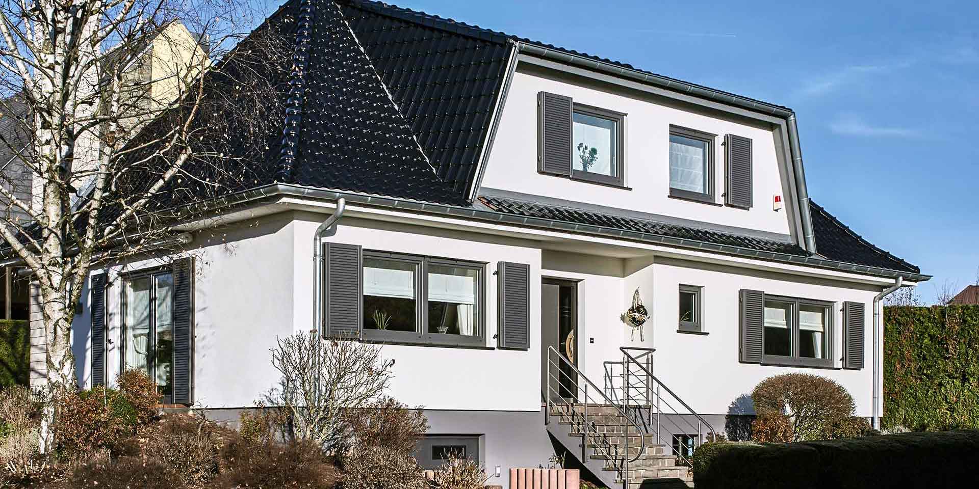 Weißes Einfamilienhaus mit grauen modernen Klappläden, einer grauen Coplaning Haustür und einer grauen Garage mit Lichtausschnitten.