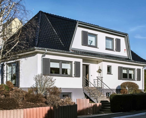 Weißes Einfamilienhaus mit grauen modernen Klappläden, einer grauen Coplaning Haustür und einer grauen Garage mit Lichtausschnitten.