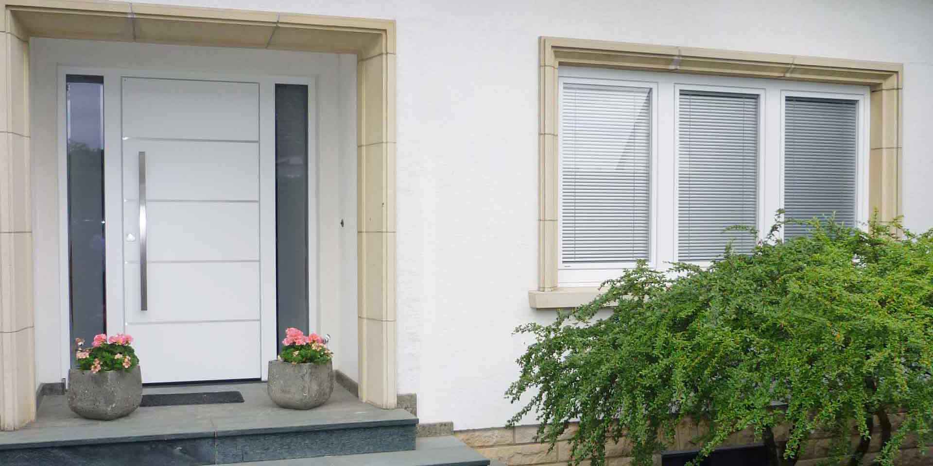 Nahaufnahme auf den Eingangsbereich des weißen Einfamilienhauses mit Blick auf die neue moderne weiße Coplaning Haustür und rechts davon drei weißen Kunststoff Coplaning Fenster.