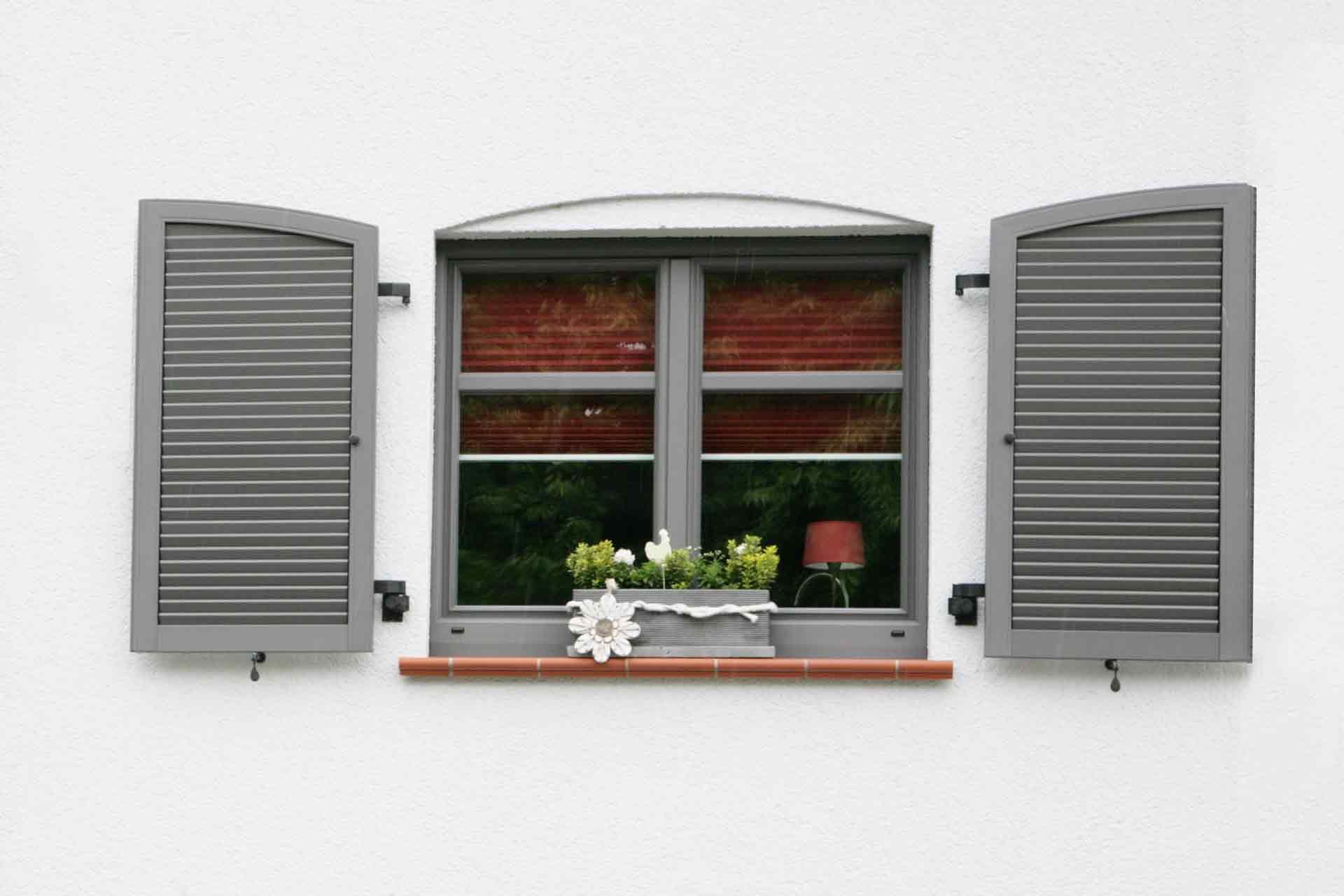 Nahaufnahme eines weiß- grauen Einfamilienhauses mit Blick auf ein graues Coplaning Alu Fenster mit grauen Klappläden.