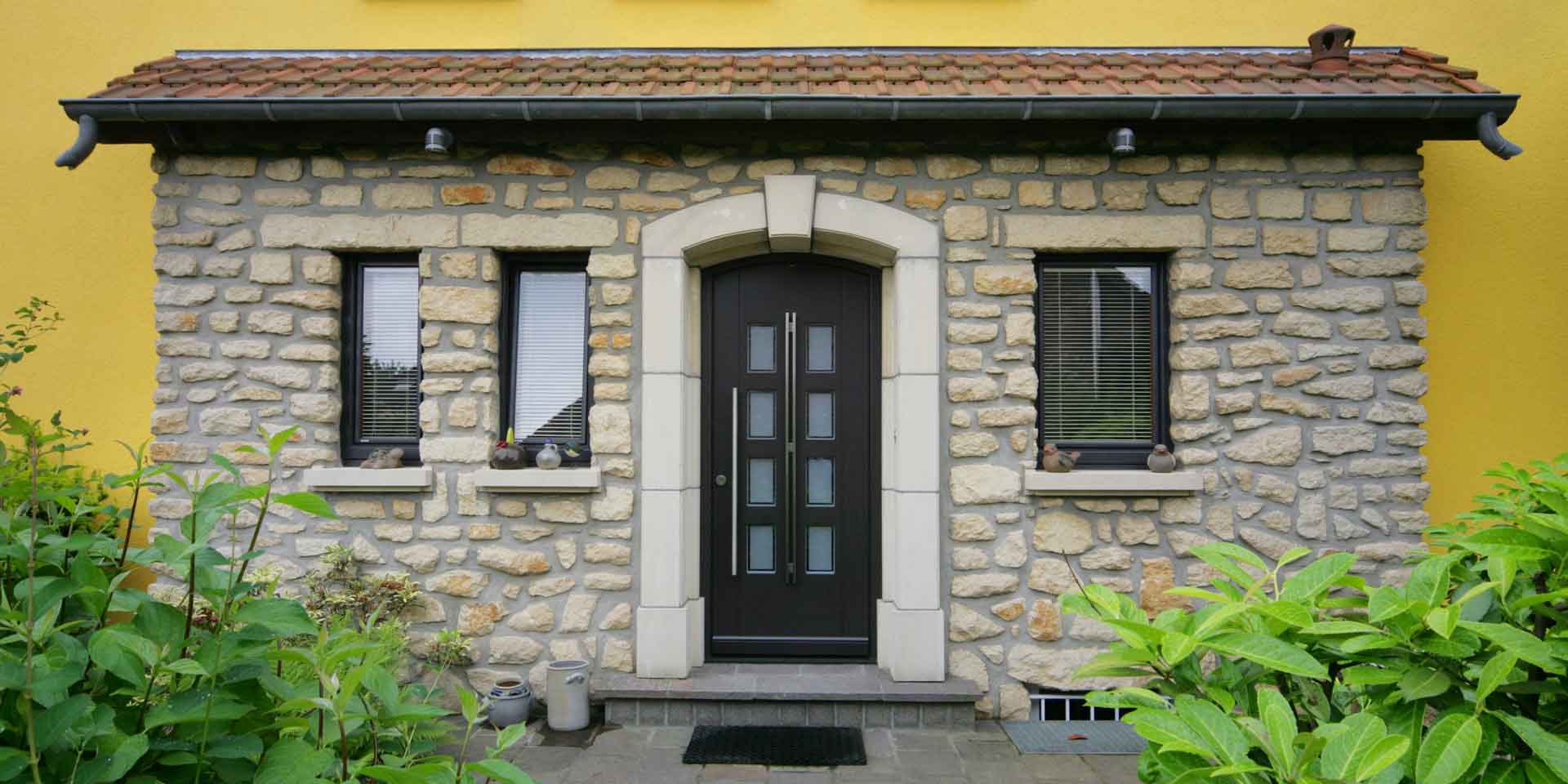 Nahaufnahme des Eingangsbereis eines gelben Einfamilienhauses mit einer rustikal braunen Coplaning Haustür und mit braunen Coplaning Holz Jalousiefenstern.