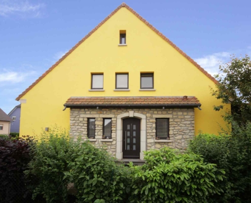 Gelbes Einfamilienhaus mit brauner Coplaning Haustür und braunen Coplaning Holz Jalousiefenster.