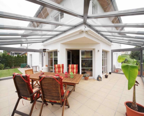 Weißes Einfamilienhaus mit Nahaufnahme der Rückseite des Hauses mit Blick auf eine Coplaning Veranda Glasschiebewand mit einer grauen Aluminium Glasüberdachung mit Terassenüberdachung um die Ecke.