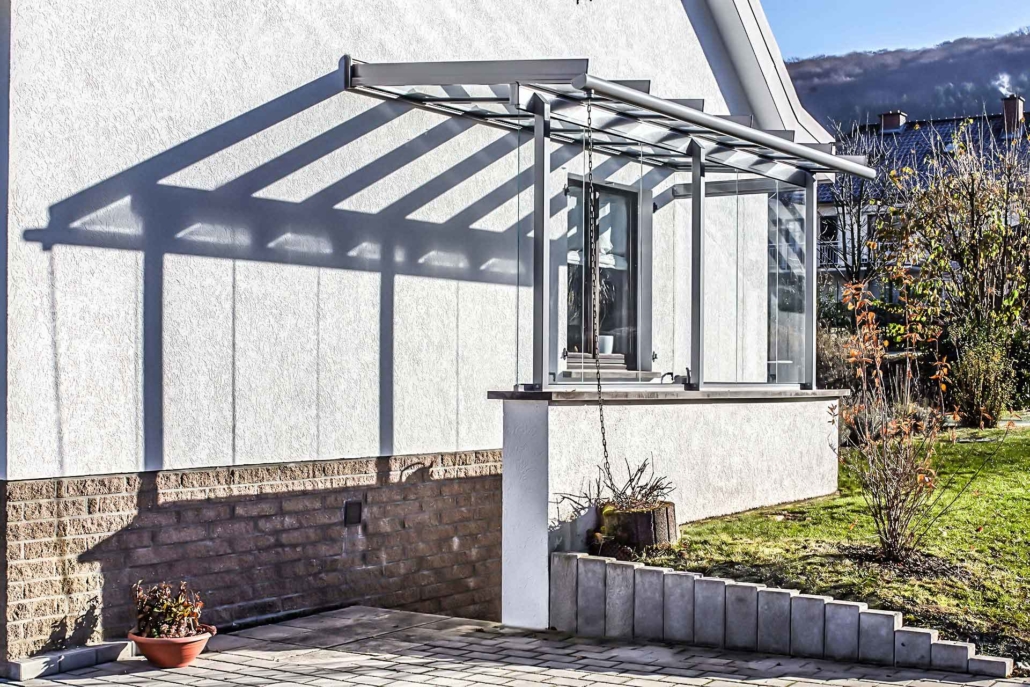 Weißes Einfamilienhaus mit einer Coplaning Veranda Überdachung aus Glas und Aluminium mit grauem Witterungsschutz.