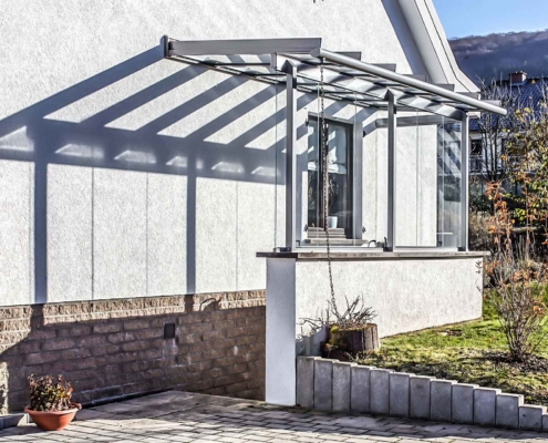 Weißes Einfamilienhaus mit einer Coplaning Veranda Überdachung aus Glas und Aluminium mit grauem Witterungsschutz.
