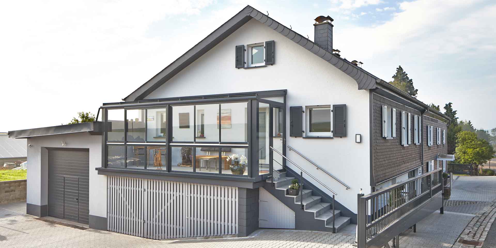 Weißes Einfamilienhaus mit einer Coplaning Veranda Überdachung aus Aluminium, grauem Balkon mit Treppenbereich und einer Verglasung mit Schiebefenster und integrierter Beleuchtung.