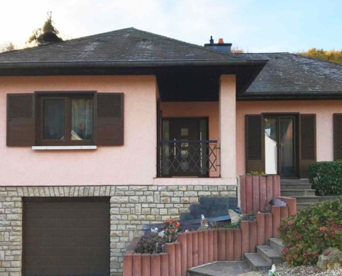 Vorderseite eines leicht pinken Einfamilienhauses mit brauner modernen Coplaning Haustür und braunen Fensterläden.