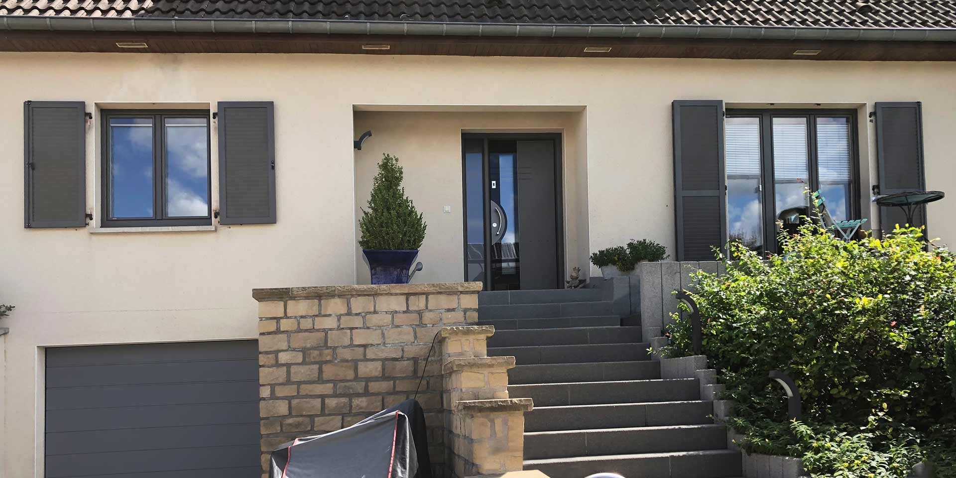 Nahaufnahme auf den Eingangsbereich des weißen Einfamilienhauses mit moderner grauer Coplaning Haustür und grauen Coplaning Holzfenster und Klappläden links und rechts von der Coplaning Haustür.
