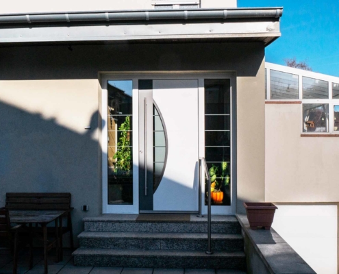 Eingang eines grauen Einfamilienhauses mit moderner weißer Coplaning Haustür.
