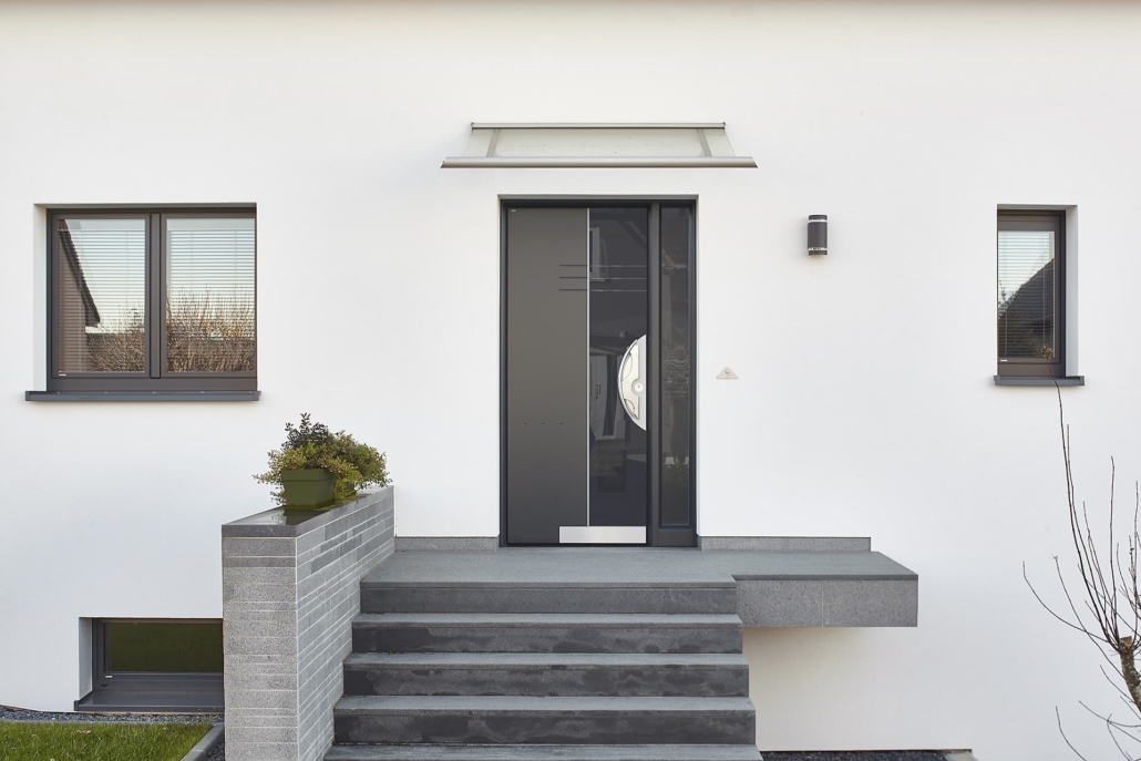 Nahaufnahme des Eingangsbereichs eines weißen Einfamilienhauses mit Blick auf die graue Coplaning Haustür mit rechts und links grauen Coplaning Alu Fenster.