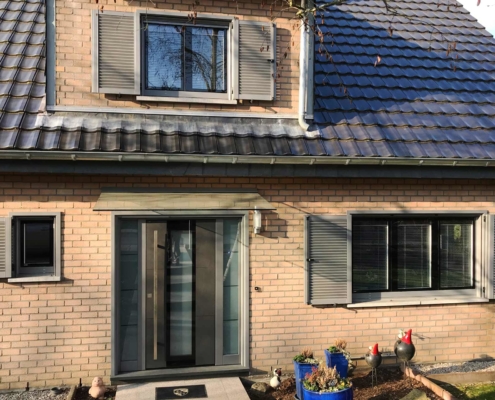 Nahaufnahme des Eingangsbereichs eines Einfamilienhauses mit Steinfassade mit Blick auf eine graue Coplaning Keramik Haustür und jeweils links und rechts davon ein graues Coplaning Holzfenster mit passenden Klappläden.