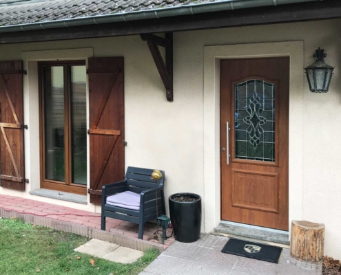 Nahaufnahme vom Eingangsbereich des beigen Einfamilienhauses. Man sieht die Coplaning Haustür aus Holz und links davon passende braune Coplaning Alu Fenster.