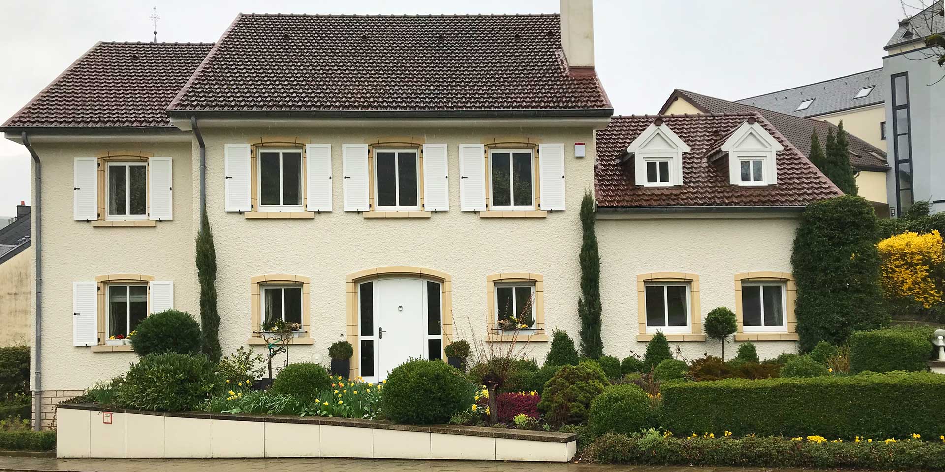 Gelbliches Einfamilienhaus mit moderner weißer Coplaning Haustür und weiße Coplaning Holzfenster mit weißen Klappläden.